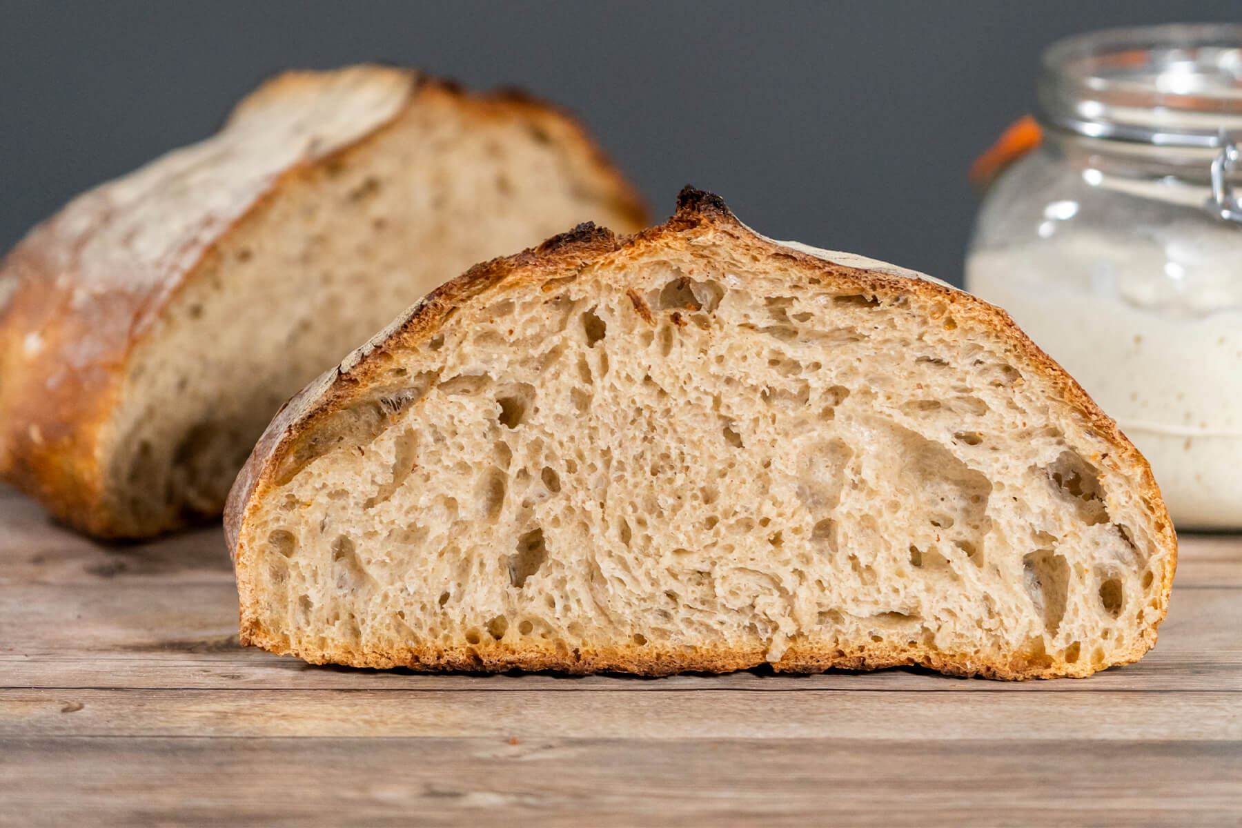 Angeschnittenes Sauerteigbrot mit offener Krume und kräftiger Kruste auf Holz, im Hintergrund ein Bügelglas mit hellem Anstellgut
