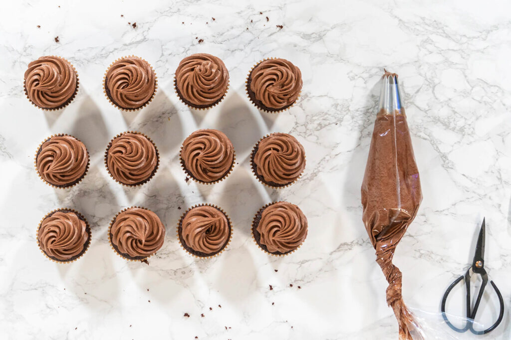 Zwölf Schokoladen-Cupcakes mit aufgespritzten Rosetten aus hellbrauner Schoko-Buttercreme von oben, daneben Spritzbeutel und Schere