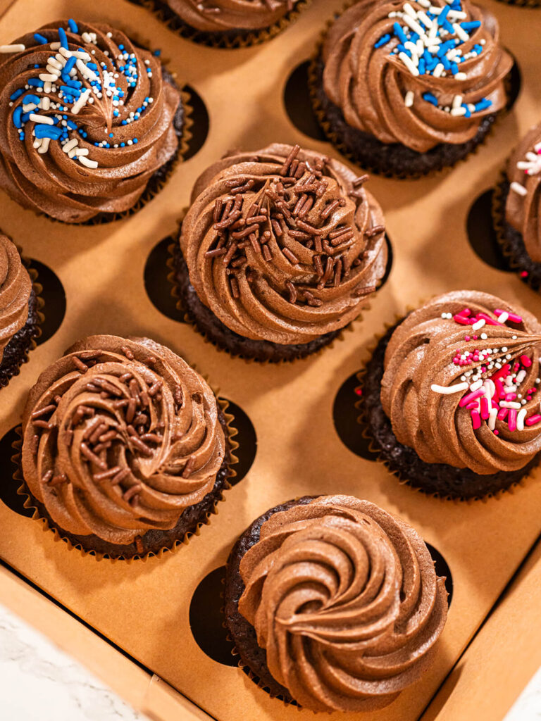 Fertige Schokoladen-Cupcakes mit Buttercreme-Rosetten in einer Transportbox, bestreut mit blau-weißen, rosa-weißen und Schokostreuseln