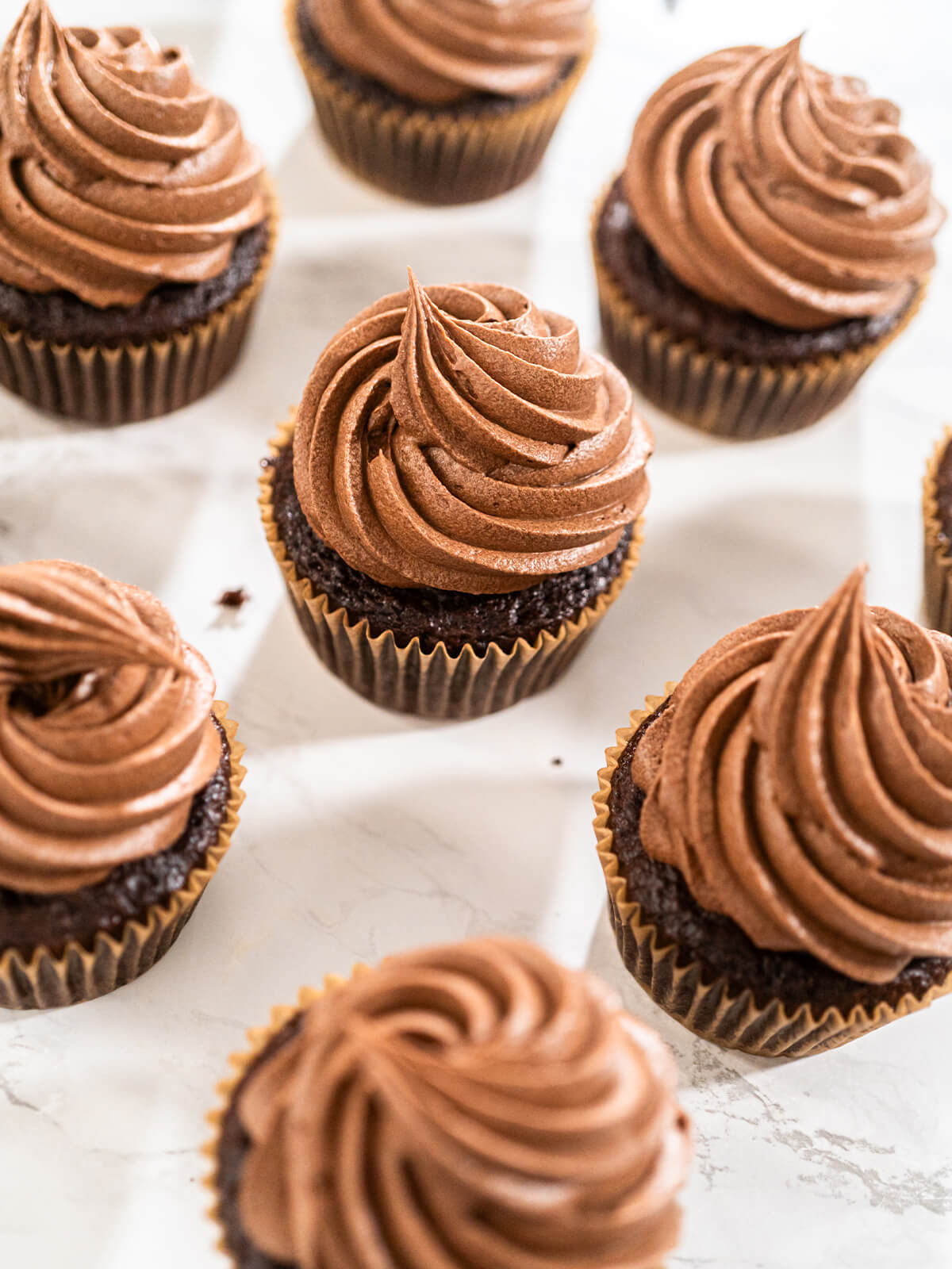 Mehrere Schokoladen-Cupcakes mit gedrehten Rosetten aus hellbrauner Schoko-Buttercreme, schräg von oben auf hellem Untergrund