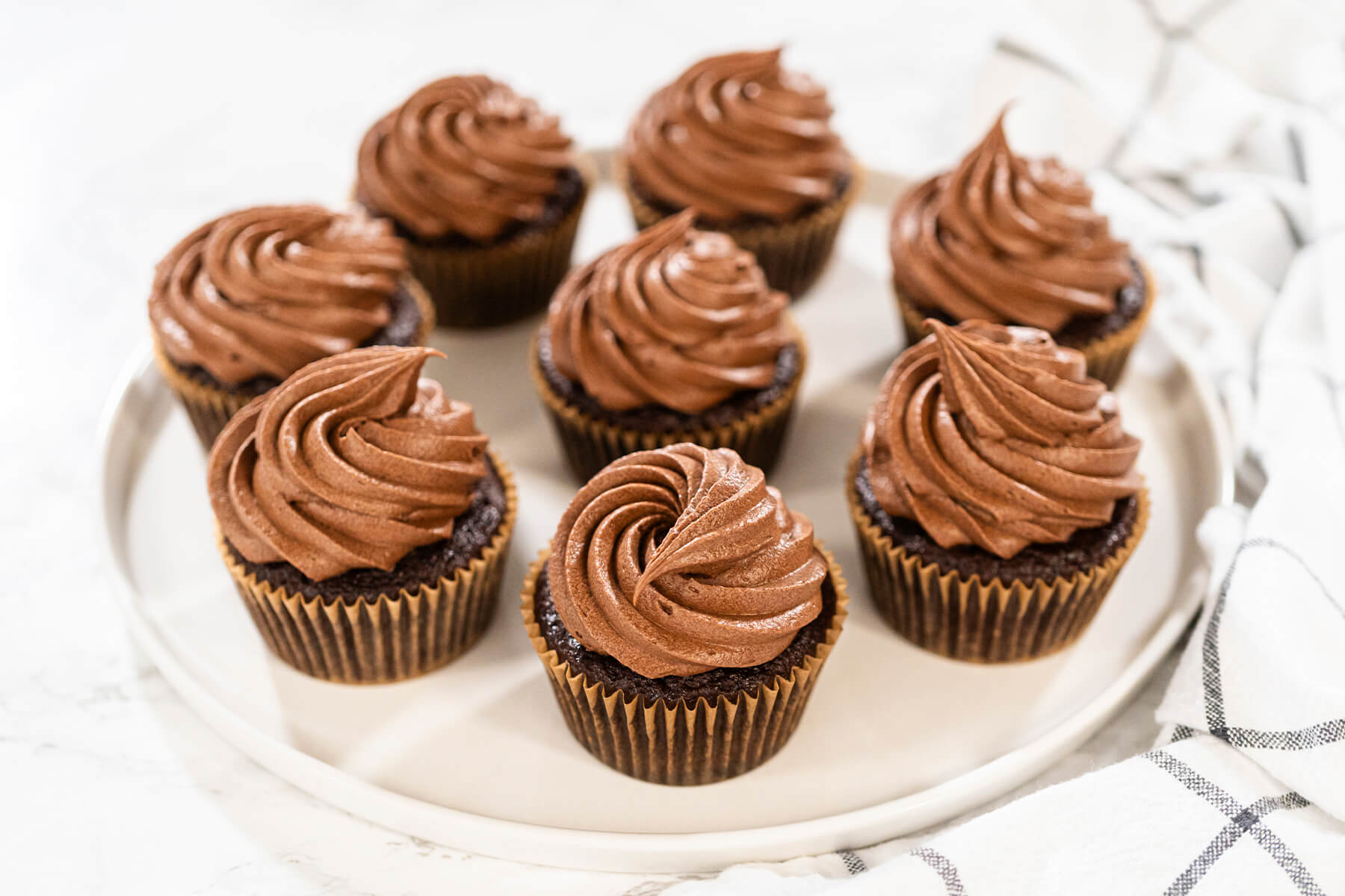 Sieben Schokoladen-Cupcakes mit Buttercreme-Rosetten auf einer hellen runden Platte, daneben ein kariertes Küchentuch