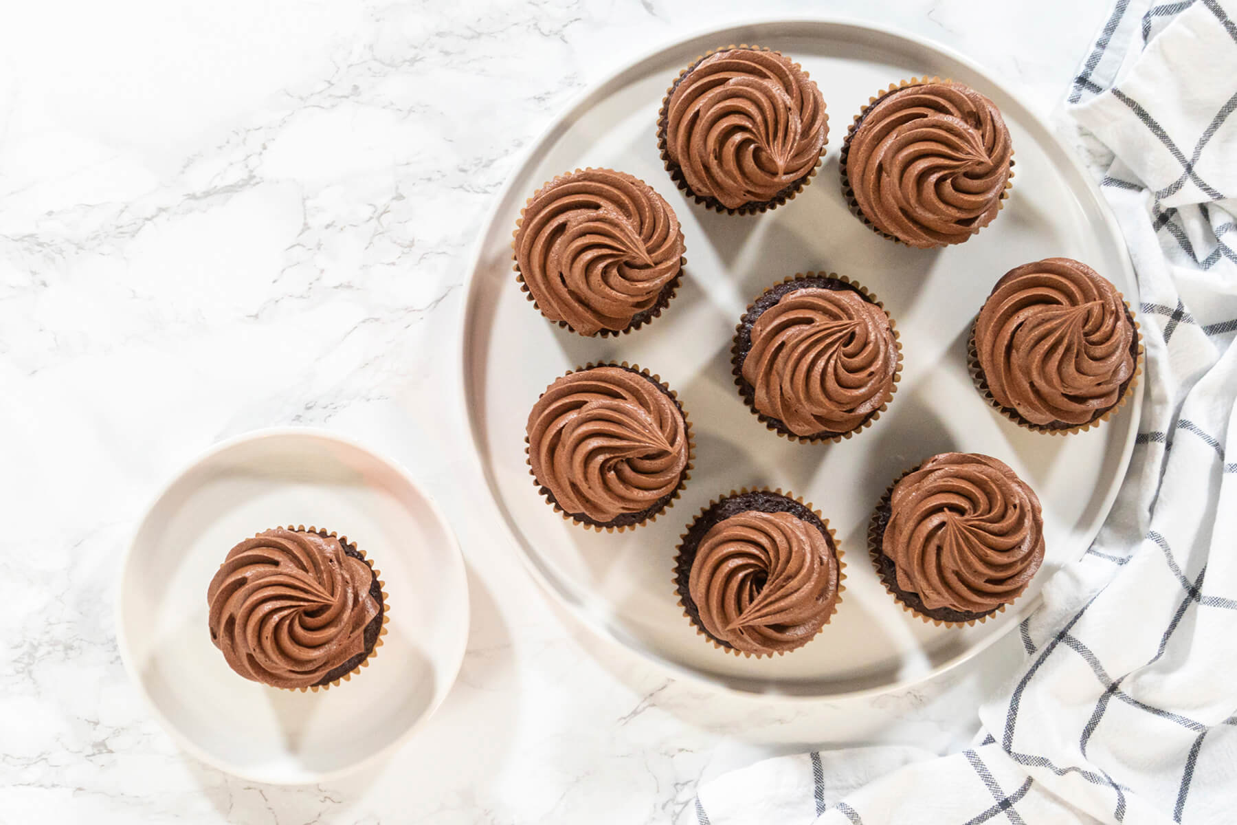 Neun Schokoladen-Cupcakes mit Buttercreme-Rosetten auf einer runden Servierplatte, ein einzelner Cupcake auf einem kleinen Teller daneben