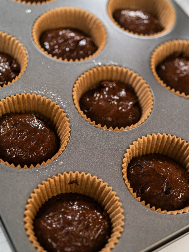 Dicker glänzender Schokoladenteig als kleine Hügel in braunen Papierförmchen einer Muffinform, von oben fotografiert