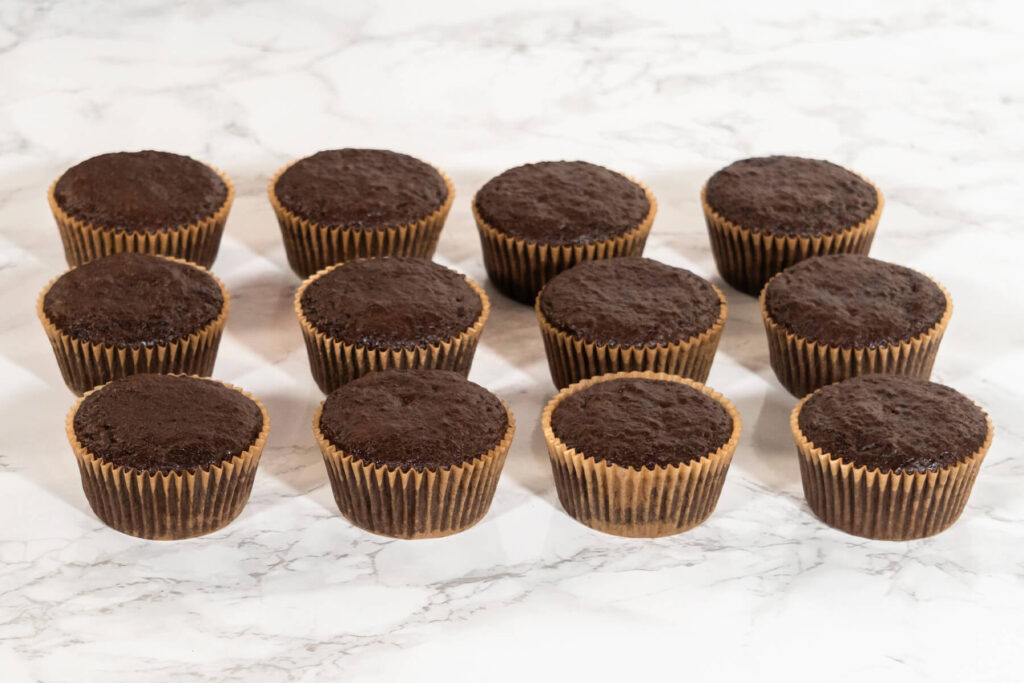 Zwölf ausgekühlte Schokoladen-Cupcakes mit flachen Oberflächen in braunen Papierförmchen in drei Reihen auf heller Marmorplatte