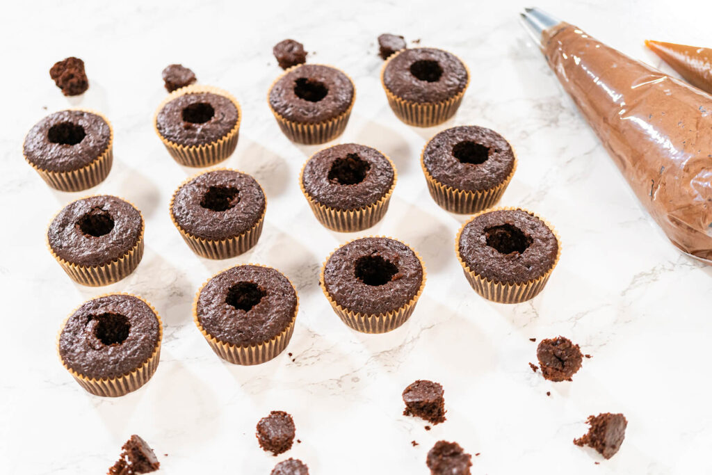 Zwölf Schokoladen-Cupcakes mit ausgestochener Mulde in der Mitte, daneben die ausgestochenen Teigstücke und zwei gefüllte Spritzbeutel