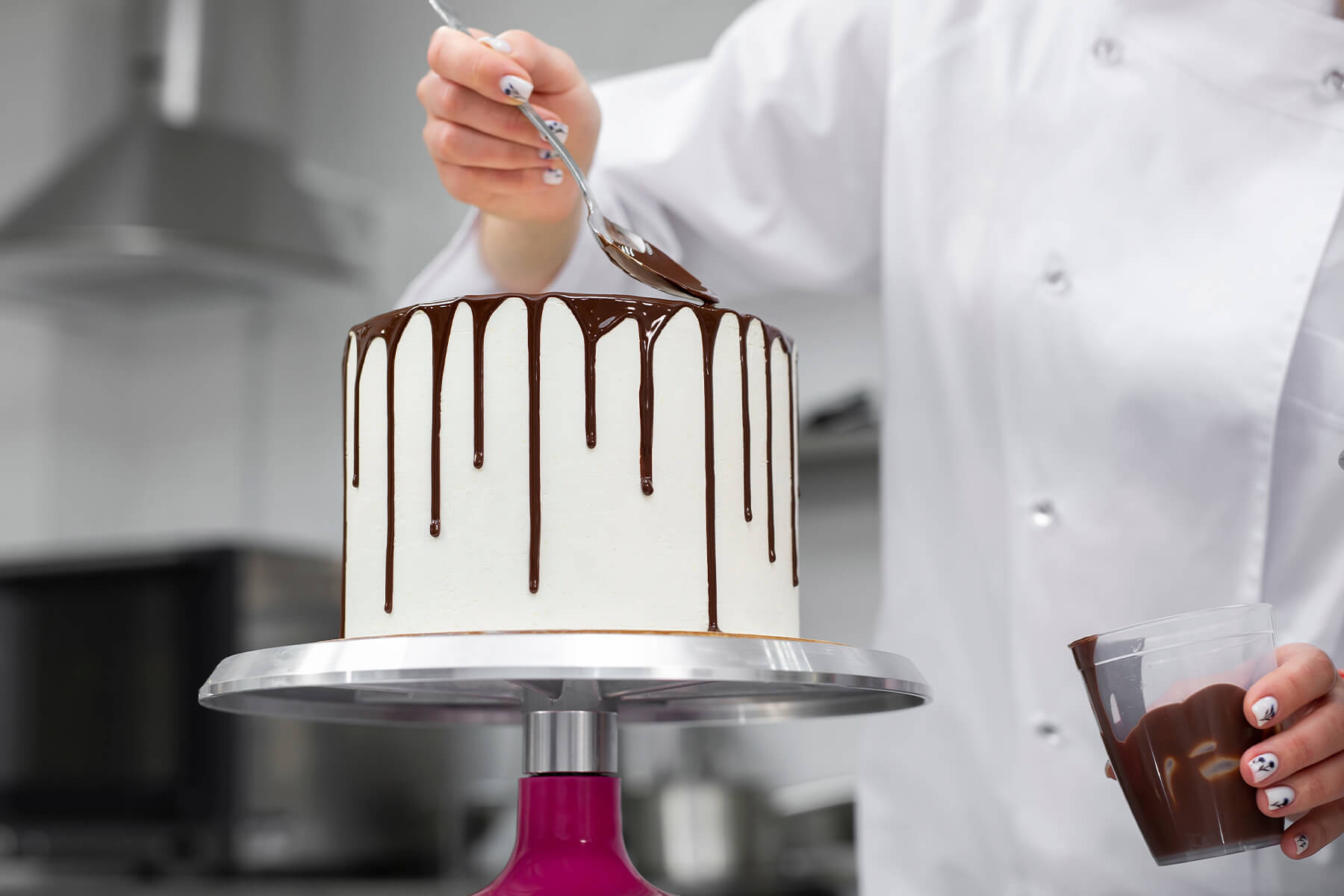 Ganache läuft in unterschiedlich langen Nasen über die Kante einer weißen Torte, in der zweiten Hand steht der Becher mit der restlichen Masse