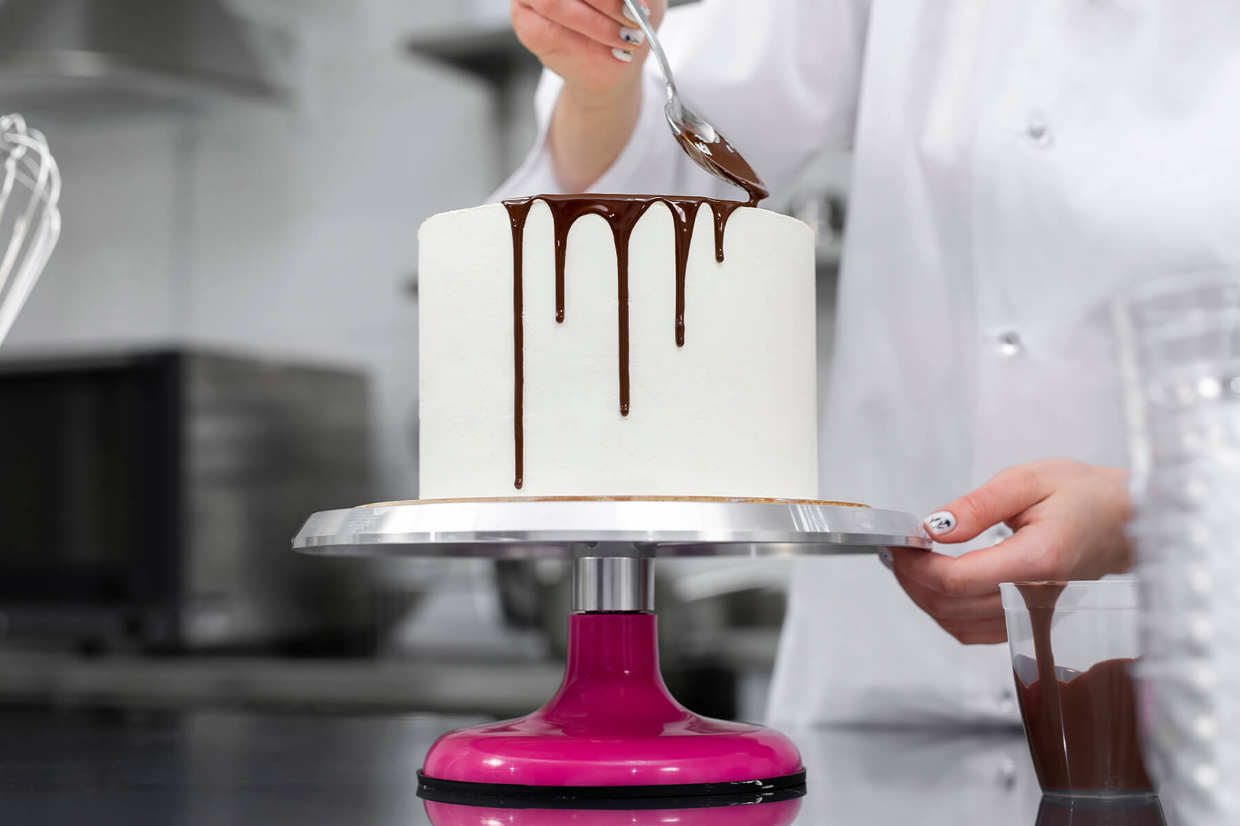 Ganache wird mit einem Löffel über die obere Kante einer weiß eingestrichenen Torte geschoben, die zweite Hand dreht den Drehteller
