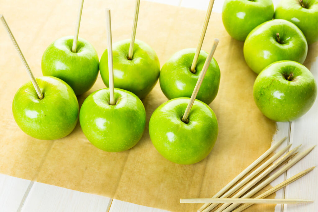 Sieben grüne Granny-Smith-Äpfel ohne Glasur auf braunem Backpapier, in jedem steckt von oben mittig ein heller Holzspieß; rechts liegen weitere ganze grüne Äpfel und ein Bündel unbenutzter Holzspieße auf einem weiß gestrichenen Holztisch
