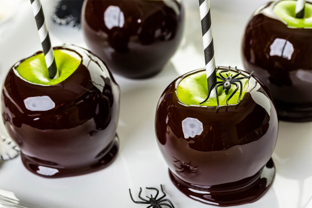Vier fast schwarze Liebesäpfel auf einem weißen Servierteller, die Glasur hochglänzend und an den Rändern rotbraun durchscheinend, unten jeweils zu einem flachen Fuß ausgelaufen; auf einem Apfel sitzt eine schwarze Plastikspinne neben dem schwarz-weiß gestreiften Halm, eine zweite liegt auf dem Teller