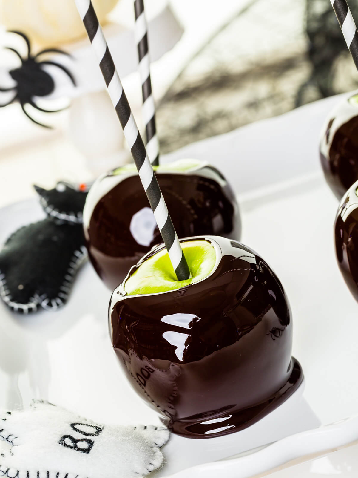 Zwei fast schwarze Liebesäpfel mit schwarz-weiß gestreiften Papierstrohhalmen auf einer weißen Servierplatte in hellem Tageslicht, die Glasur läuft an beiden zu einem breiten glänzenden Fuß aus; links im Bild eine schwarze Filzkatze und ein weißer Filzanhänger mit der Aufschrift BOO, oben rund um die Halme jeweils ein Ring hellgrüner Apfelschale