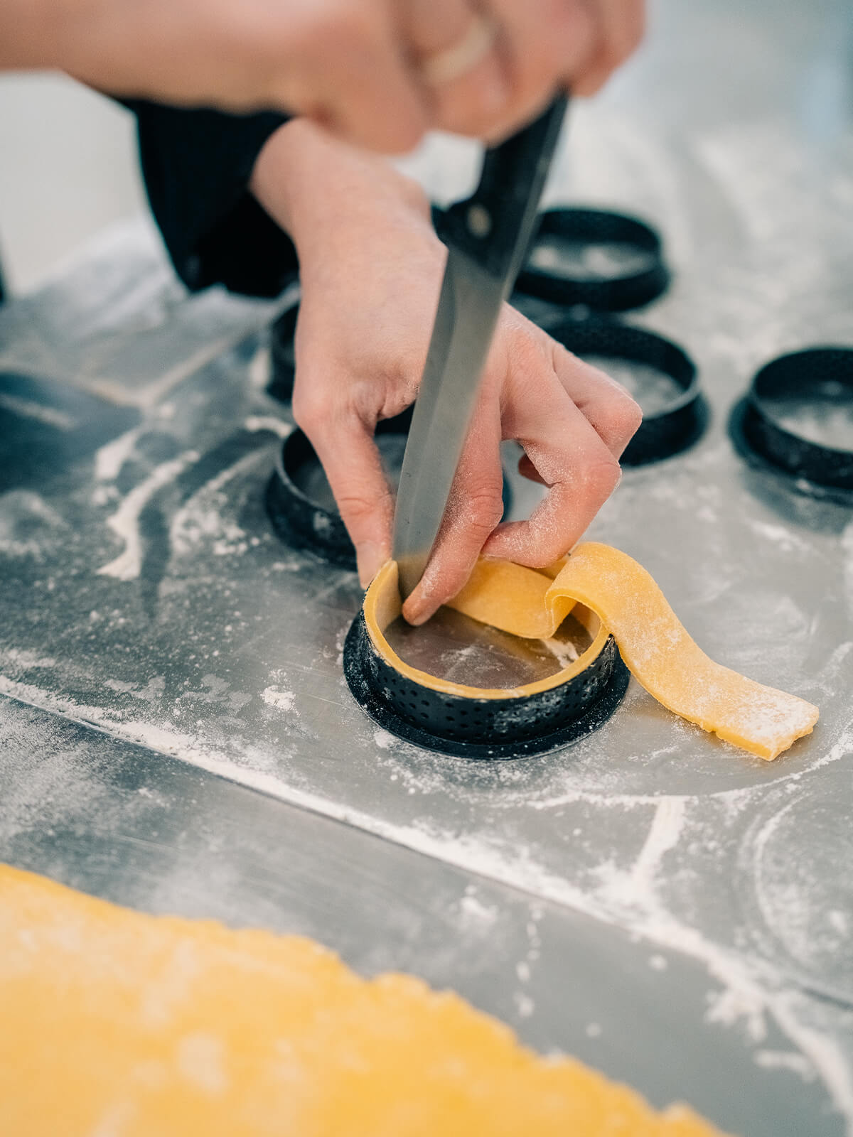 Ein Messer wird flach über die obere Kante eines perforierten Tartelettrings geführt und trennt den überstehenden Teig ab