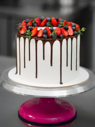 Hohe weiße Torte mit dunklem Schokoladenrand und einem Kranz aus halbierten Erdbeeren, Heidelbeeren und kleinen grünen Zweigen