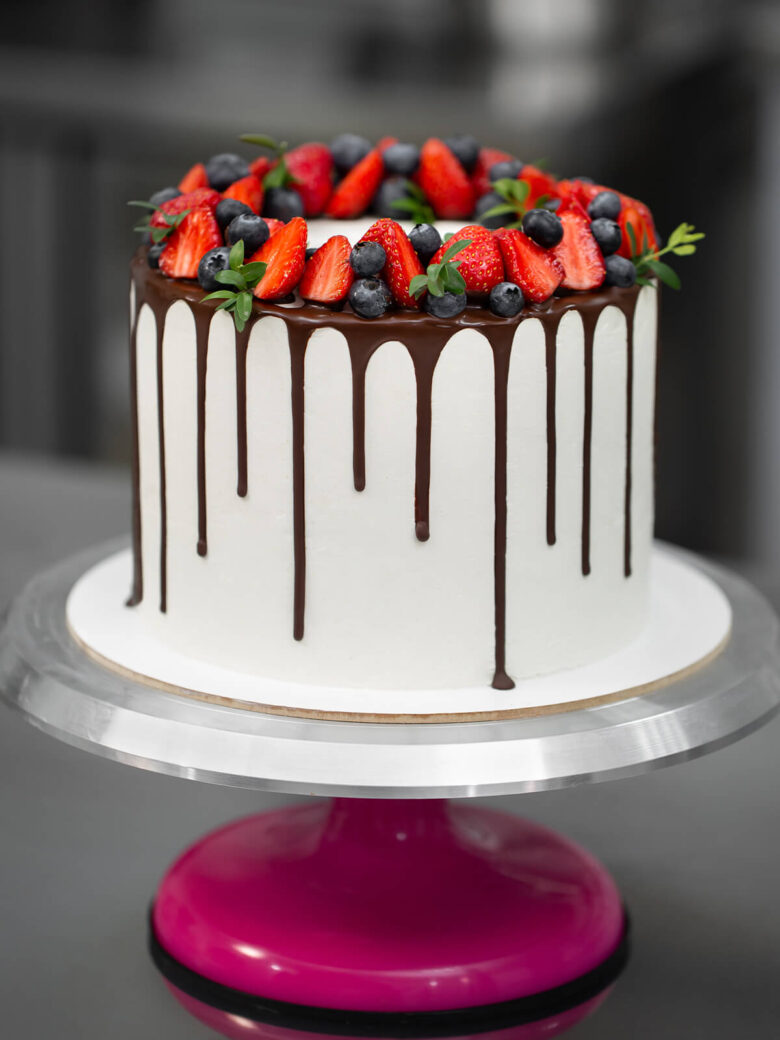 Hohe weiße Torte mit dunklem Schokoladenrand und einem Kranz aus halbierten Erdbeeren, Heidelbeeren und kleinen grünen Zweigen