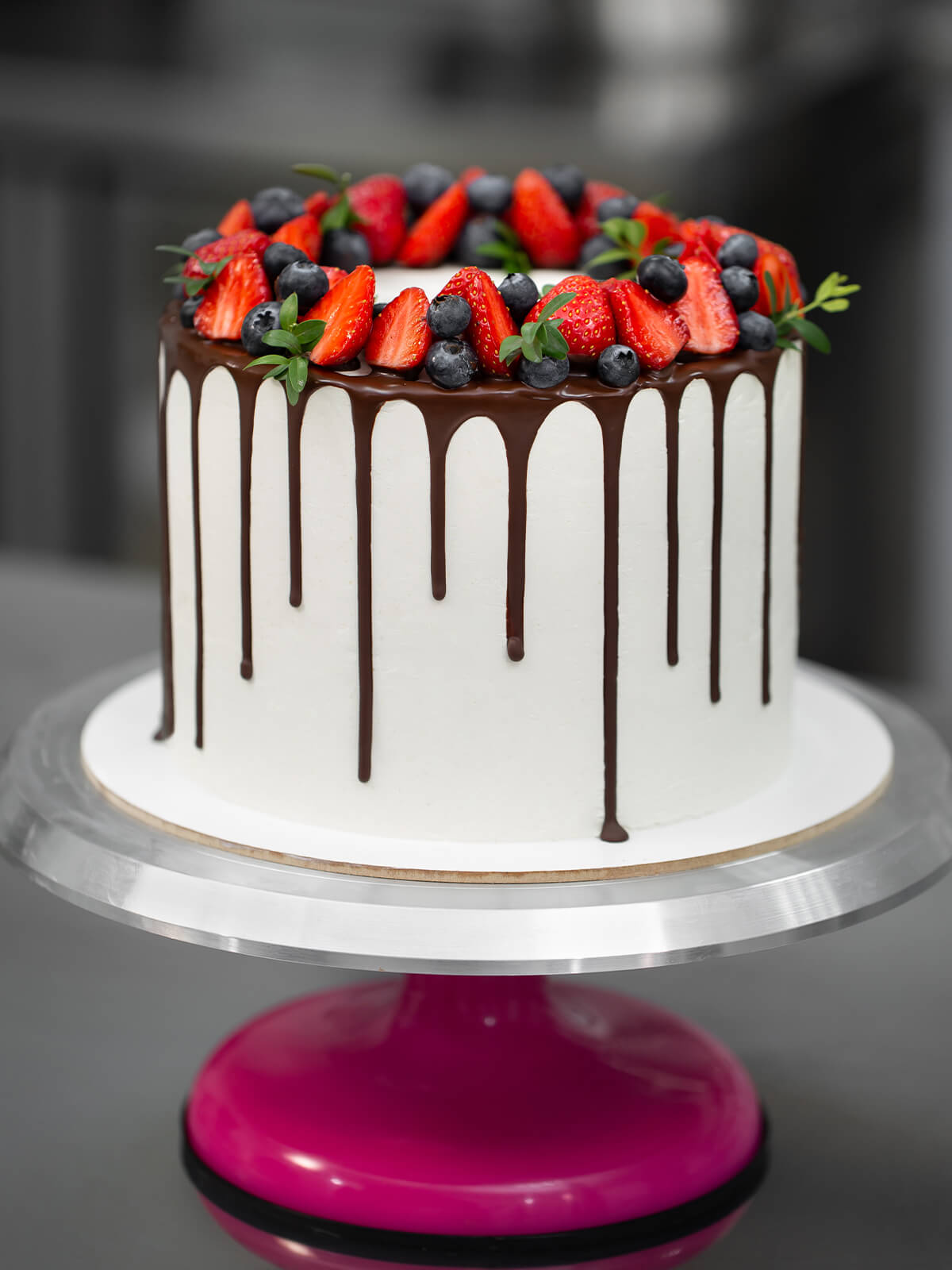 Hohe weiße Torte mit dunklem Schokoladenrand und einem Kranz aus halbierten Erdbeeren, Heidelbeeren und kleinen grünen Zweigen