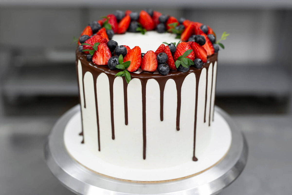 Weiße Torte mit dunklem Schokoladen-Drip und einem Kranz aus Erdbeeren, Heidelbeeren und grünen Zweigen auf dem Drehteller