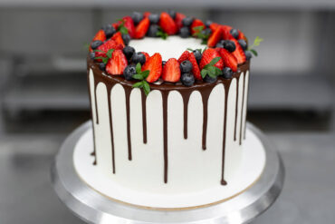 Weiße Torte mit dunklem Schokoladen-Drip und einem Kranz aus Erdbeeren, Heidelbeeren und grünen Zweigen auf dem Drehteller