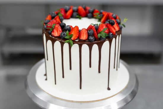 Weiße Torte mit dunklem Schokoladen-Drip und einem Kranz aus Erdbeeren, Heidelbeeren und grünen Zweigen auf dem Drehteller