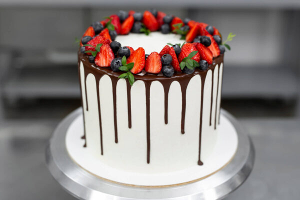 Weiße Torte mit dunklem Schokoladen-Drip und einem Kranz aus Erdbeeren, Heidelbeeren und grünen Zweigen auf dem Drehteller