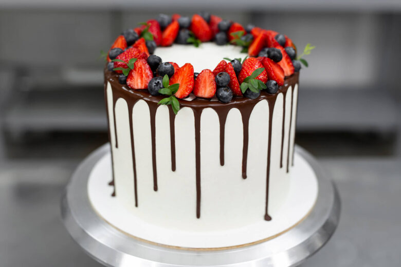 Weiße Torte mit dunklem Schokoladen-Drip und einem Kranz aus Erdbeeren, Heidelbeeren und grünen Zweigen auf dem Drehteller