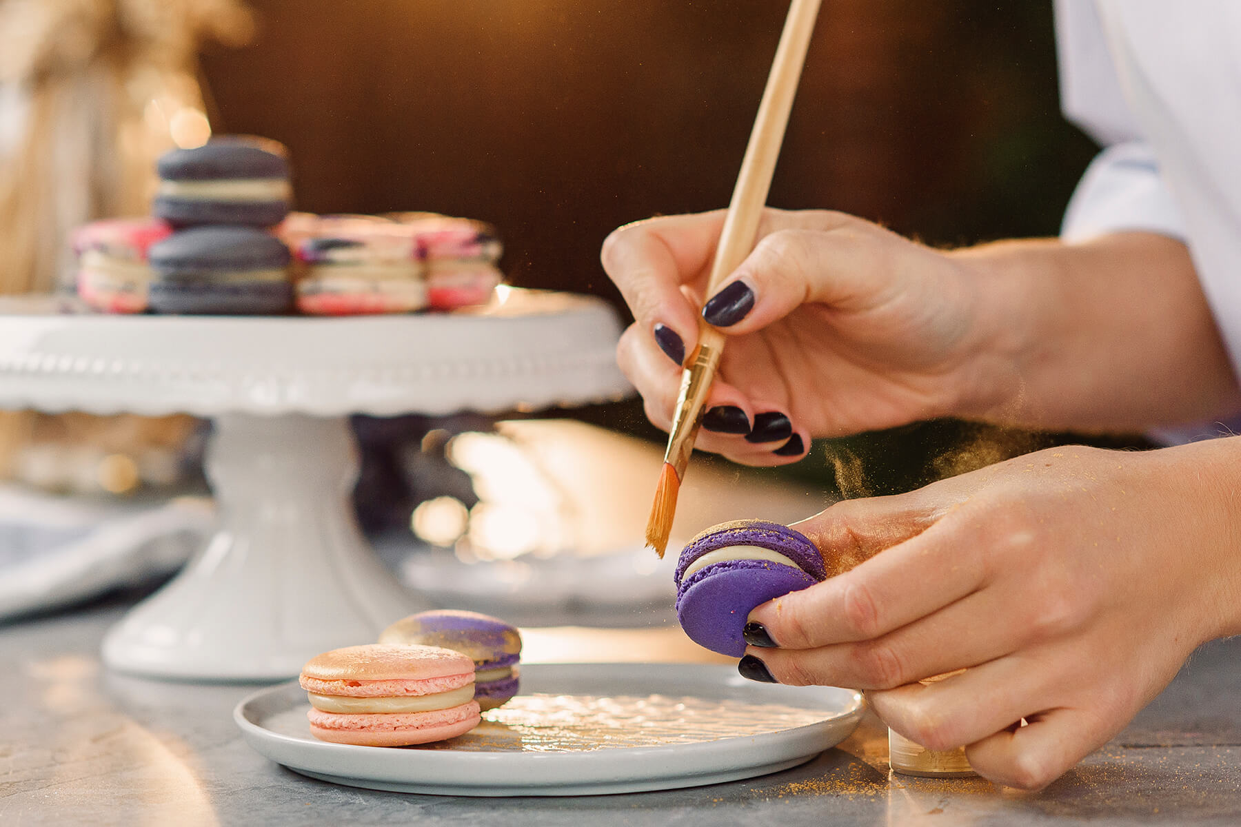 Zwei violette Macarons und ein rosafarbenes liegen auf einem Teller, im Hintergrund eine Etagere mit anthrazitfarbenen und rosa Macarons
