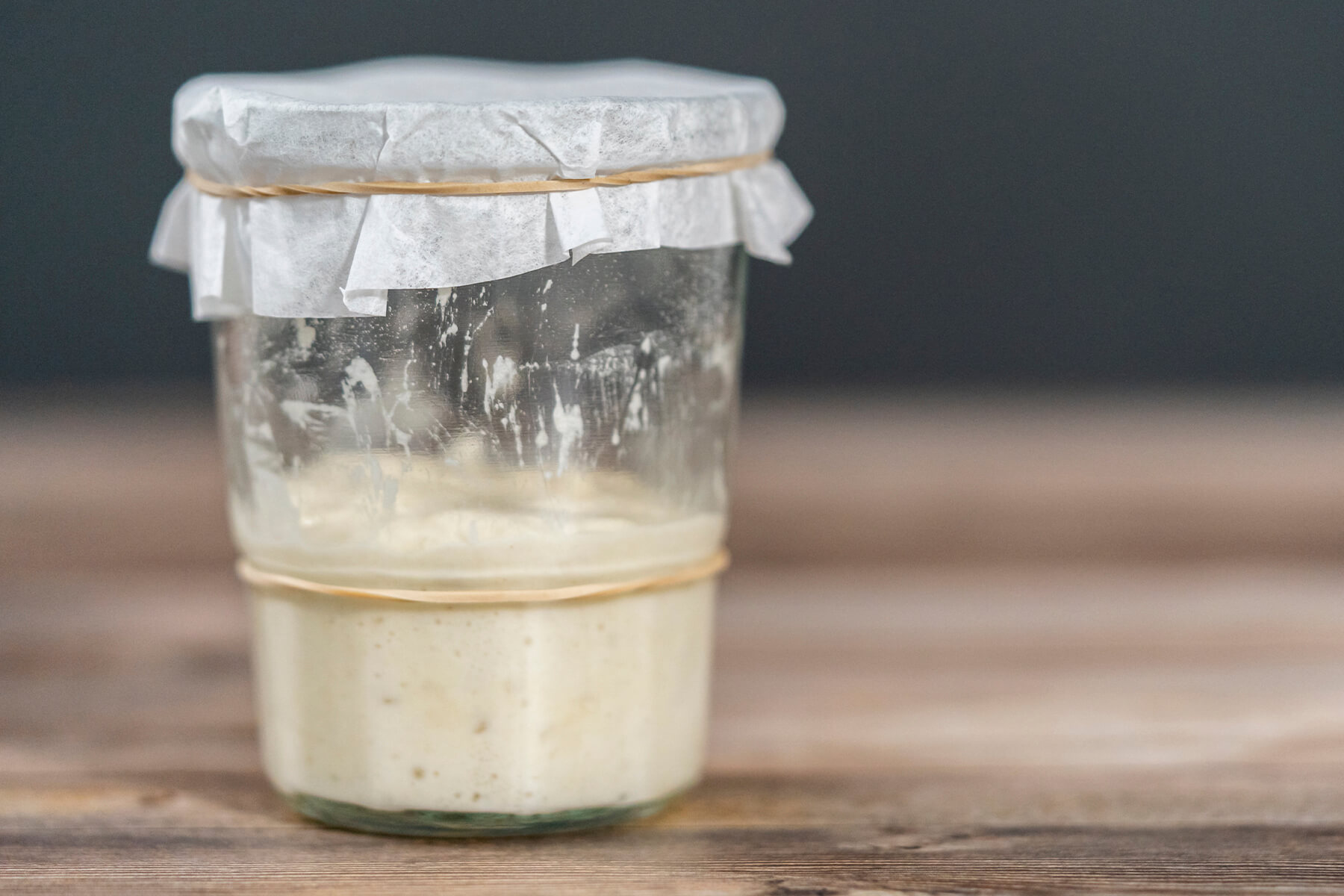 Glas mit Weizensauerteig, der wieder zusammengefallen ist, an der Glaswand stehen eingetrocknete Spuren deutlich über dem aktuellen Stand