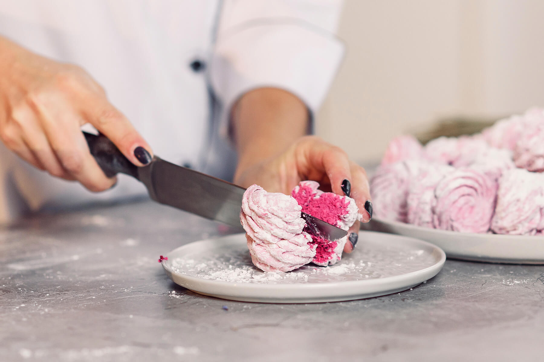 Ein Messer schneidet einen mit Puderzucker bestäubten rosa Zefir auf einem weißen Teller durch, die Schnittfläche zeigt eine feine, gleichmäßige Schaumstruktur