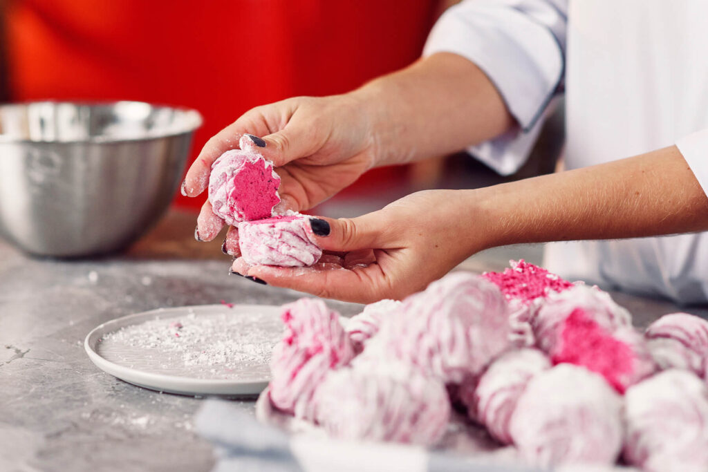 Zwei Hände setzen zwei Zefir-Hälften mit den leuchtend pinkfarbenen, feuchten Unterseiten aneinander, im Hintergrund liegen weitere Hälften auf einem Tablett