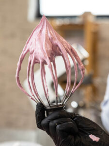 Ein Schneebesen wird aus rosa Zefirmasse gehoben, die Masse hängt in dicken Strängen an den Drähten und bildet oben eine stehende Spitze