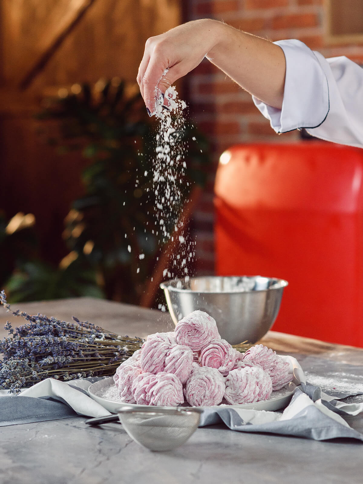Puderzucker rieselt aus einer Hand auf einen Teller mit rosa Schaumzuckergebäck, im Vordergrund liegt ein feines Sieb
