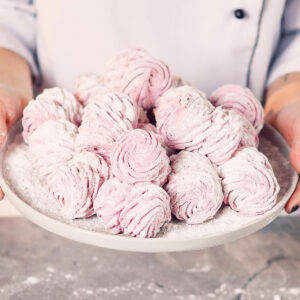 Zwei Hände halten einen grauen Teller mit rosa Zefir-Hälften, die paarweise zusammengesetzt und dick mit Puderzucker bestäubt sind