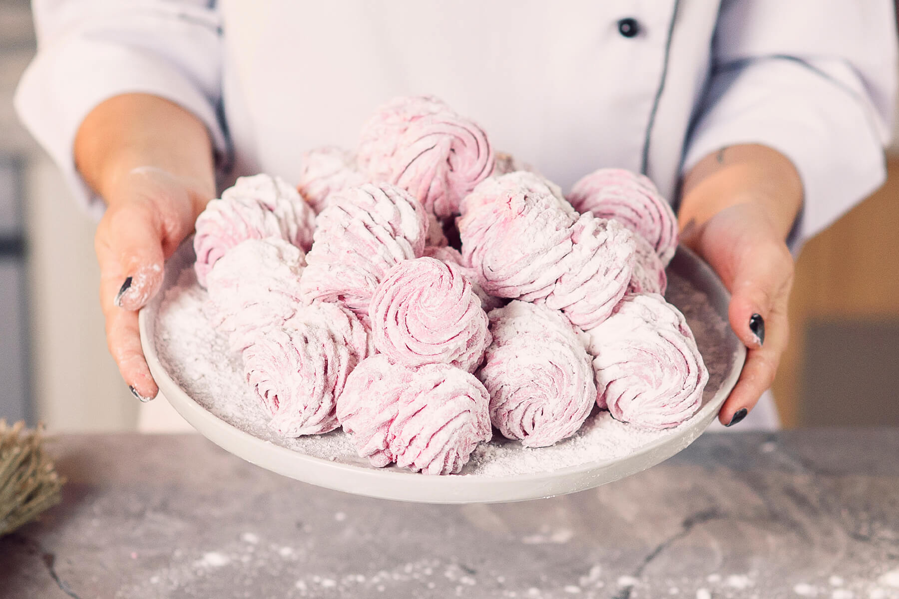 Zwei Hände halten einen grauen Teller mit rosa Zefir-Hälften, die paarweise zusammengesetzt und dick mit Puderzucker bestäubt sind