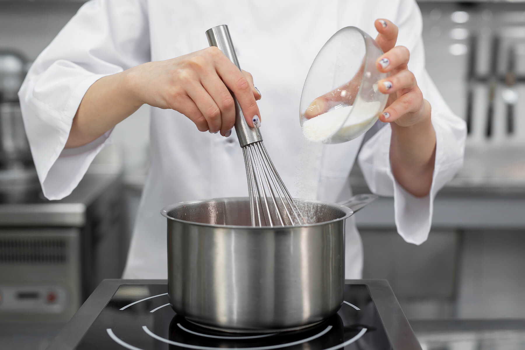 Zucker wird aus einer Glasschüssel in einen Topf gerieselt, während die andere Hand mit dem Schneebesen rührt