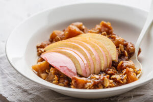 Portion Apfel-Crisp in einer weißen Schale, Apfelwürfel in dickem Sirup mit Haferflocken, darauf ein Fächer roher Apfelscheiben