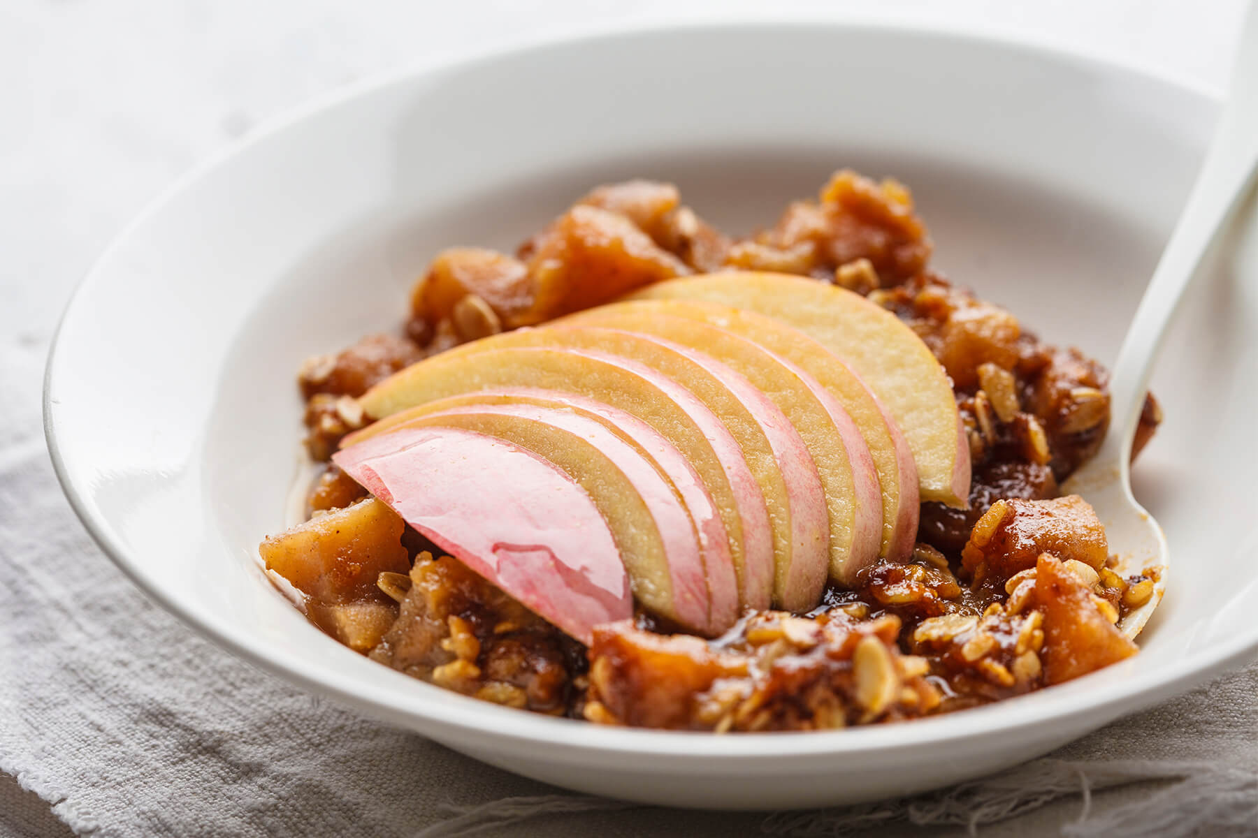 Portion Apfel-Crisp in einer weißen Schale, Apfelwürfel in dickem Sirup mit Haferflocken, darauf ein Fächer roher Apfelscheiben