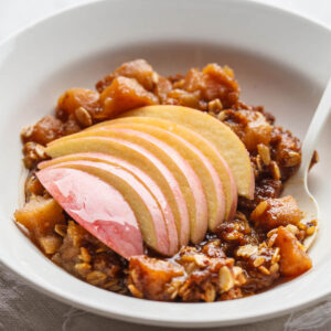 Glutenfreier Apfel-Crisp mit Haferflocken in einer weißen Schale, obenauf ein Fächer aus dünnen Apfelscheiben