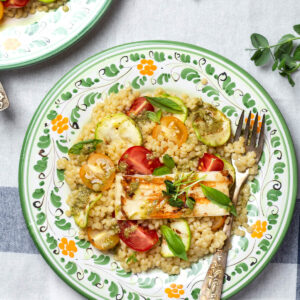 Teller mit Perlnudeln, Zucchinischeiben, roten und gelben Kirschtomaten und einer Scheibe gegrilltem Halloumi