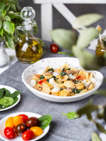 Orecchiette mit Spinat, Champignons und Mozzarella in einer weißen Schale, daneben Olivenöl und Kirschtomaten