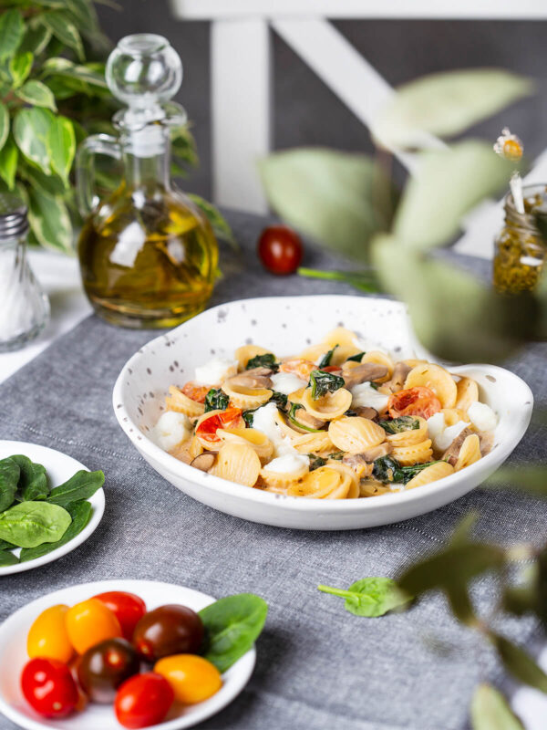 Orecchiette mit Spinat, Champignons und Mozzarella in einer weißen Schale, daneben Olivenöl und Kirschtomaten
