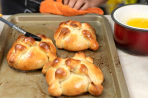 Frisch gebackene Pan de Muerto auf einem Blech werden mit zerlassener Butter bestrichen, daneben ein Topf mit flüssiger Butter