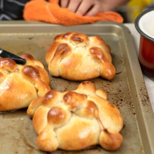 Frisch gebackene Pan de Muerto auf einem Blech werden mit zerlassener Butter bestrichen, daneben ein Topf mit flüssiger Butter