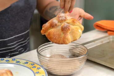 Ein gebackenes Pan de Muerto mit Teigknochen wird über einer Glasschüssel mit Zimtzucker bestreut
