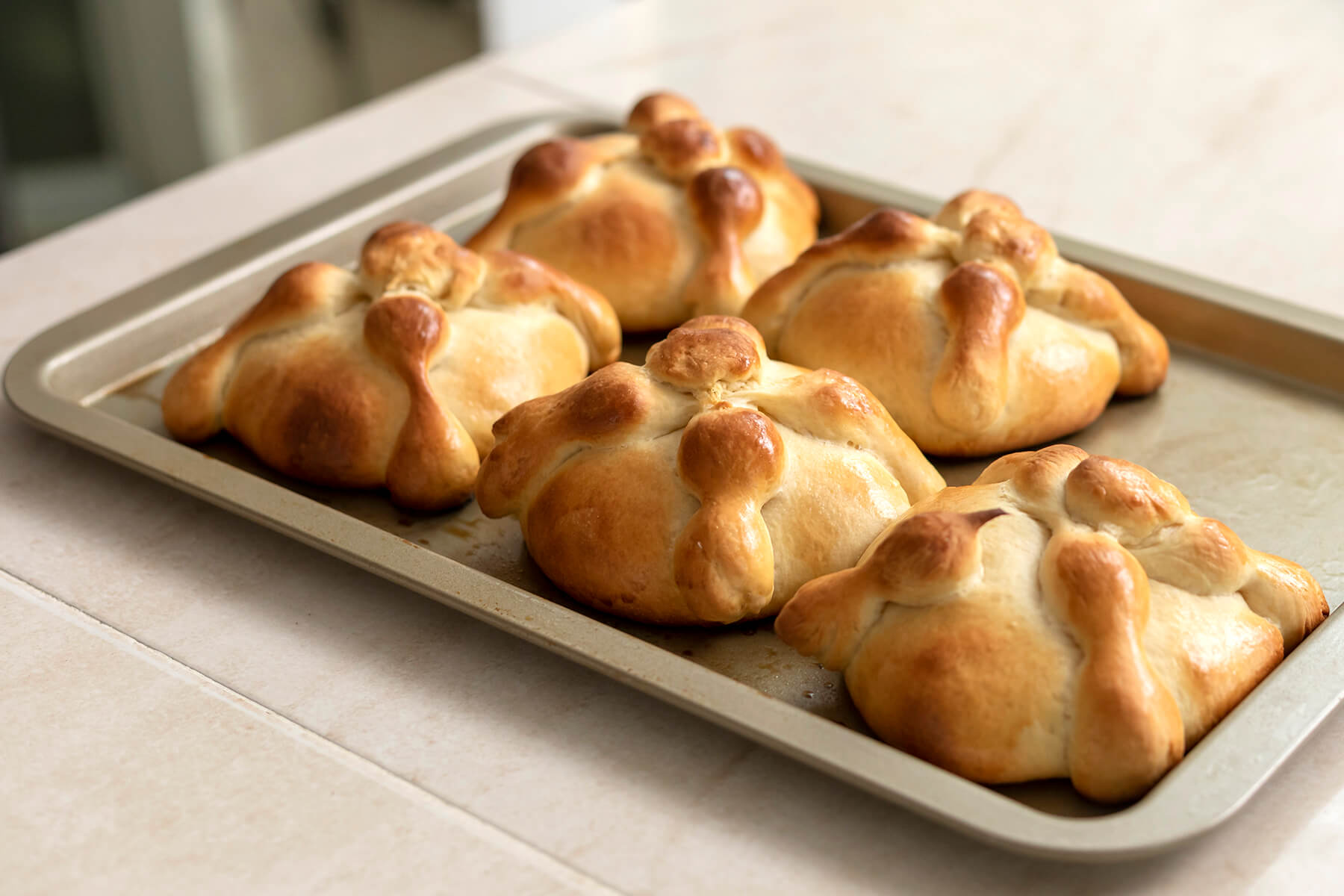 Goldbraun gebackene Pan de Muerto mit dunkel glänzenden Teigknochen, noch ohne Zucker, auf einem Backblech