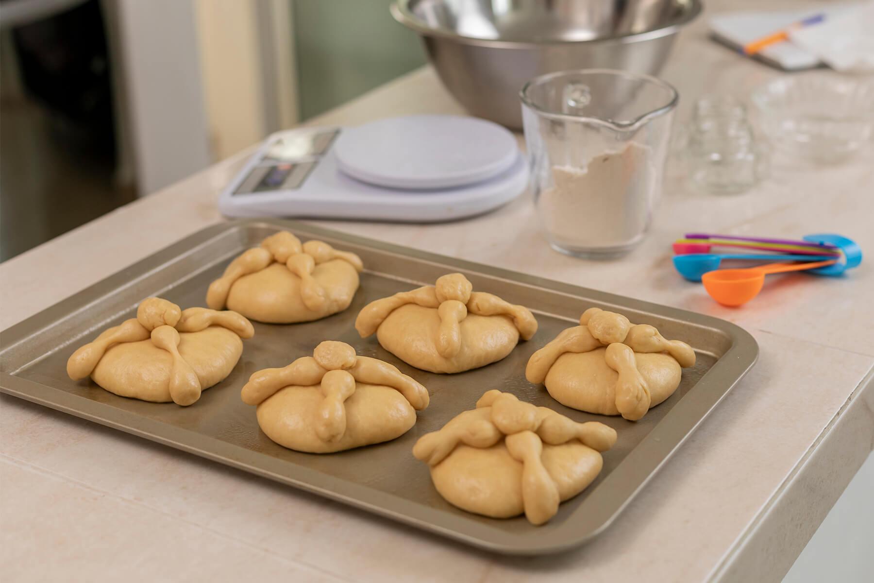Sechs ungebackene Pan de Muerto mit gekreuzten Teigknochen und kleiner Teigkugel auf einem Backblech, dahinter eine Küchenwaage und Messlöffel