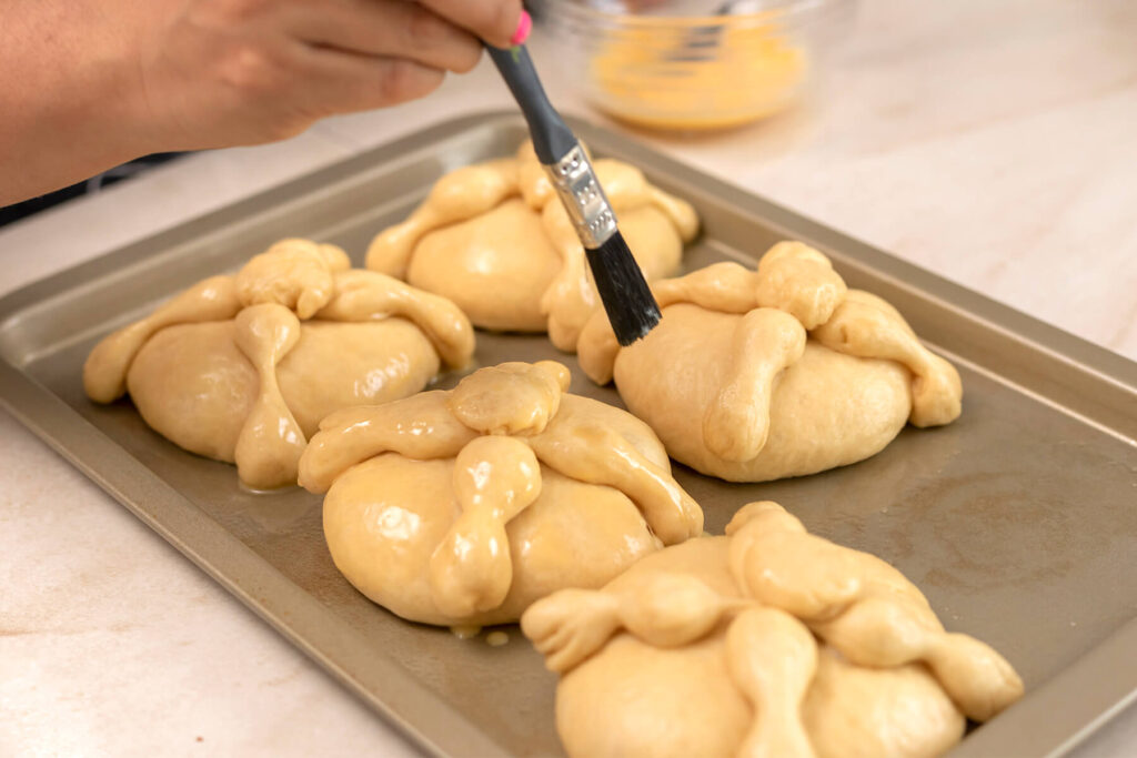 Aufgegangene Pan de Muerto werden auf dem Backblech mit verquirltem Ei eingepinselt, im Hintergrund eine Schüssel mit Ei