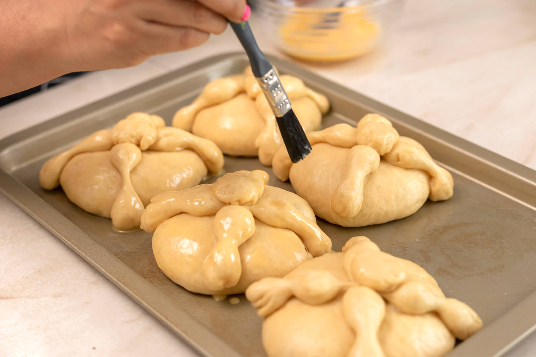 Aufgegangene Pan de Muerto werden auf dem Backblech mit verquirltem Ei eingepinselt, im Hintergrund eine Schüssel mit Ei