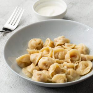 Gekochte Pelmeni mit zerlassener Butter in einem tiefen weißen Teller, daneben eine Gabel und ein Schälchen Schmand