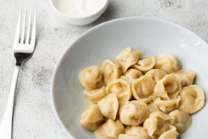 Gekochte Pelmeni mit zerlassener Butter in einem weißen Teller in Nahaufnahme, dahinter Schmand und eine Gabel