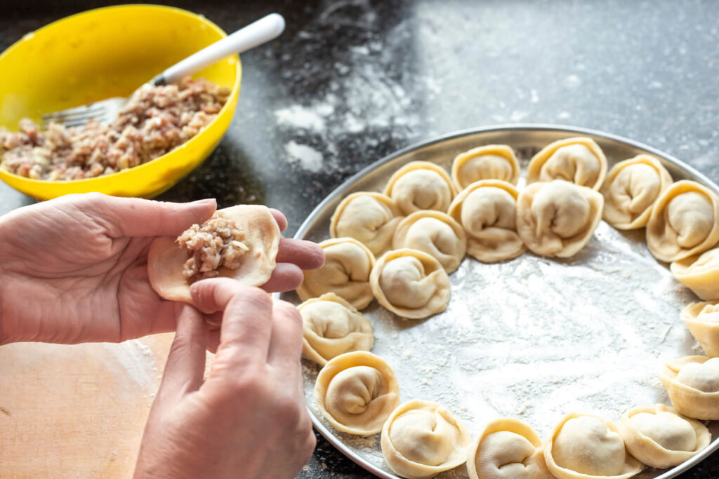 Eine Hand hält einen Teigkreis mit einem Häufchen roher Hackfleischfüllung, dahinter eine gelbe Schüssel mit Füllung und ein Blech mit fertigen Pelmeni