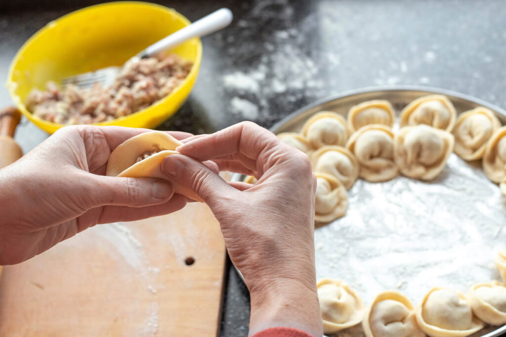 Zwei Hände drücken einen gefüllten Teigkreis zum Halbmond zusammen, rechts ein Blech mit rohen Pelmeni