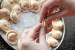 Finger drücken die beiden Enden eines gefüllten Halbmonds übereinander, darunter ein Blech mit rohen Pelmeni